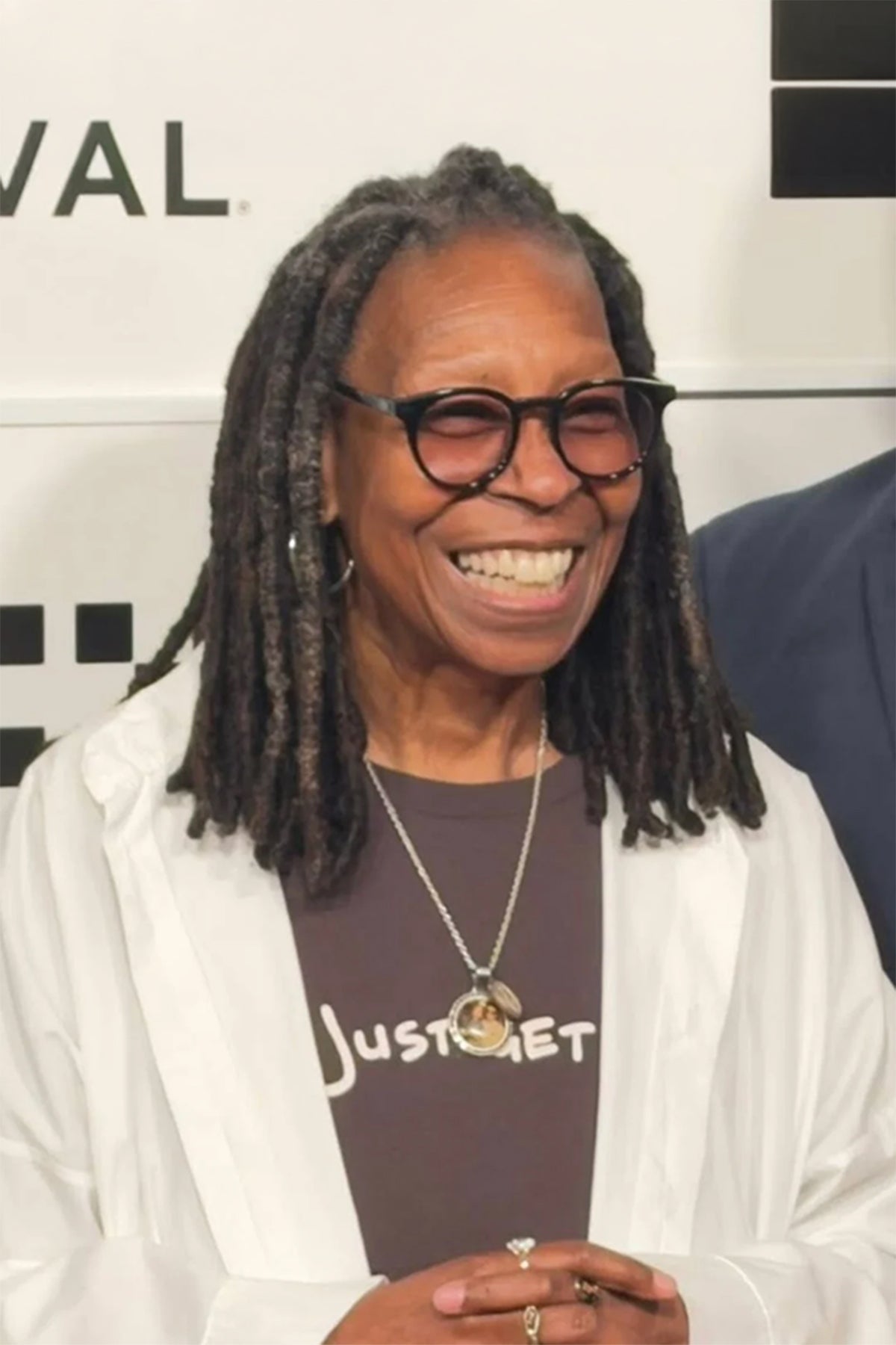 Person with dreadlocks wearing glasses and a white coat, wearing a tee shirt "Just Get Up" and smiling in front of a white background.