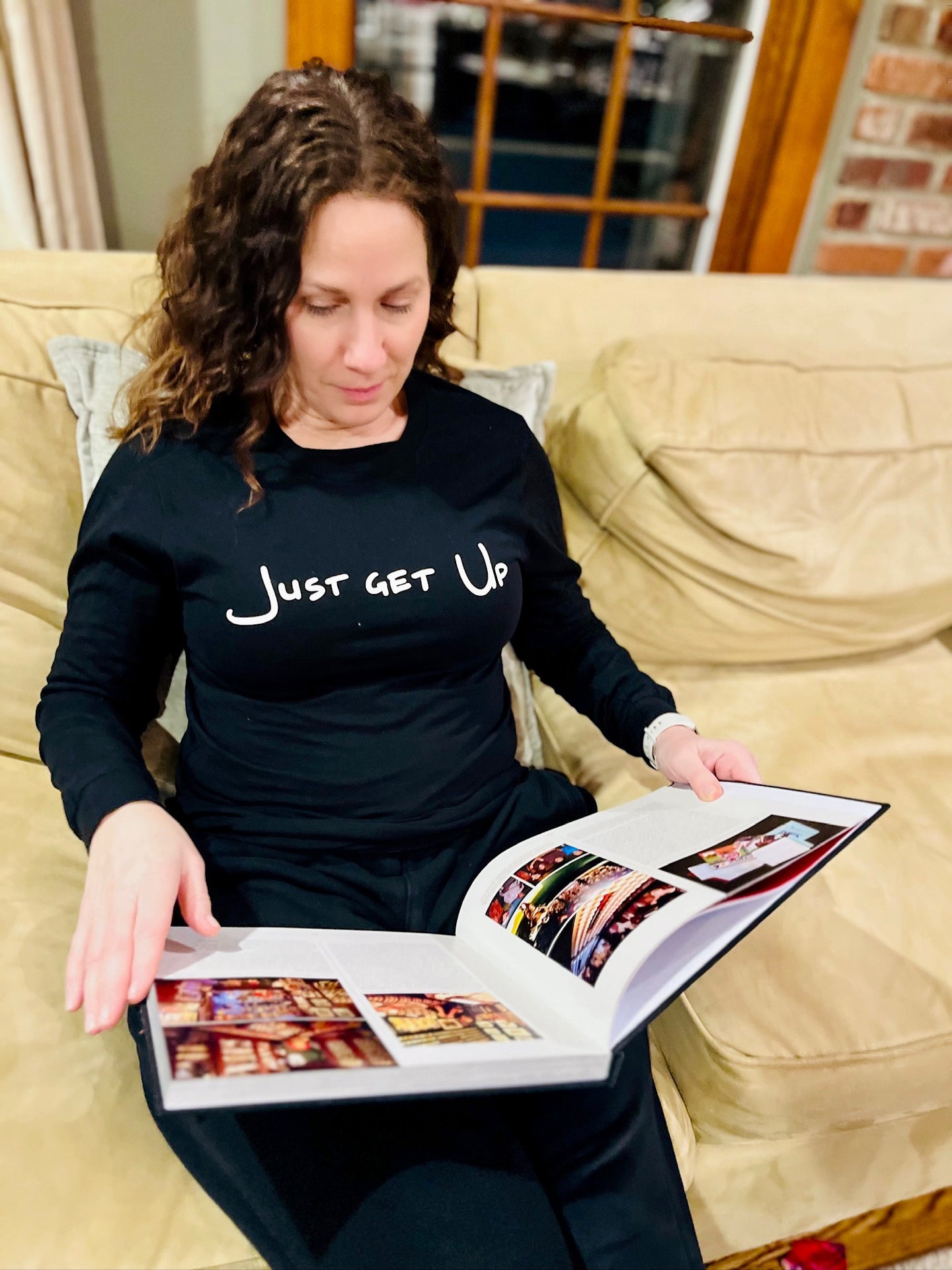 Woman sitting on a couch wearing a black shirt with 'Just Get Up' text, holding an open photo album.