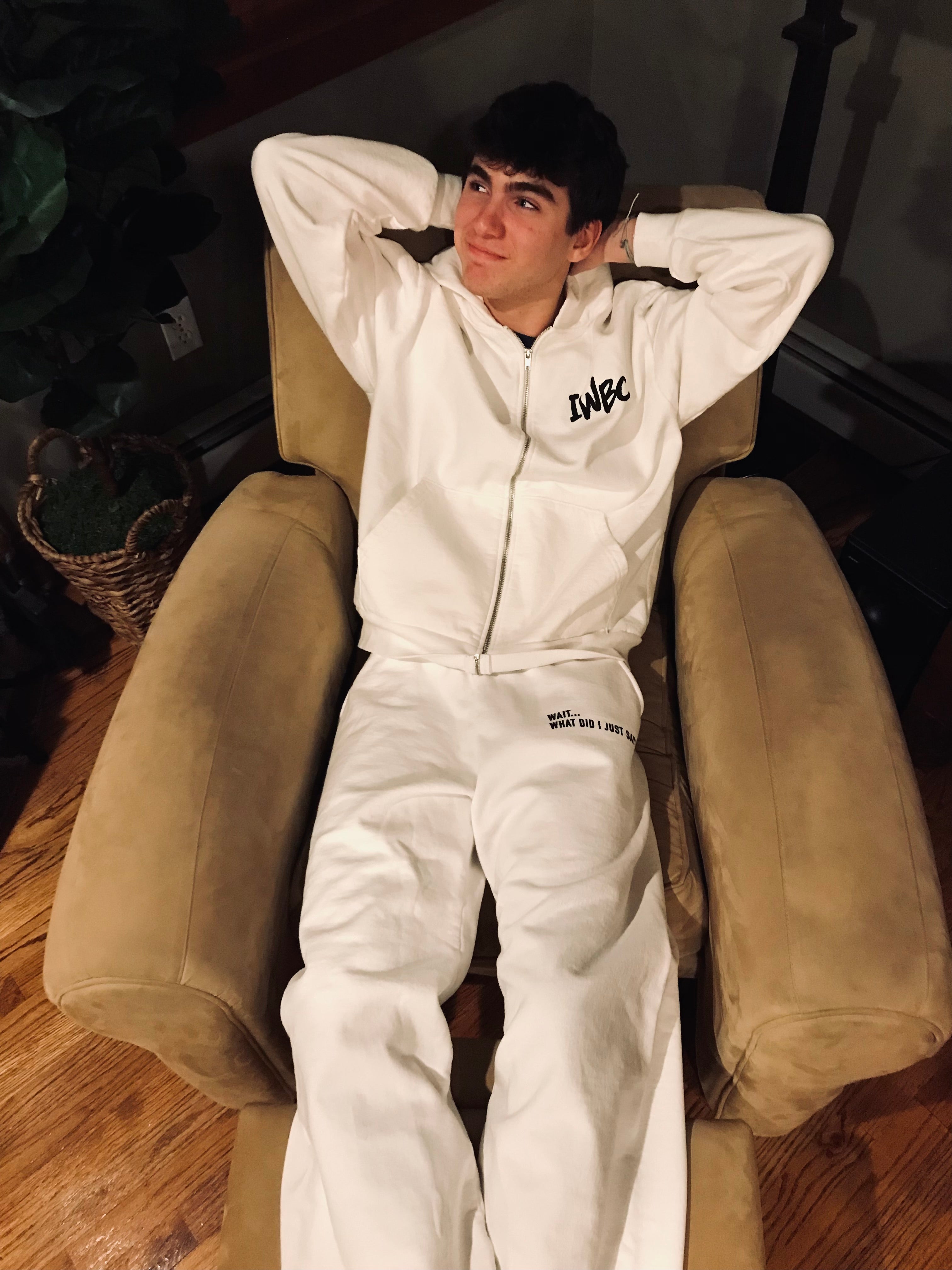 Person wearing a white hoodie and pants with text, sitting on a beige armchair.