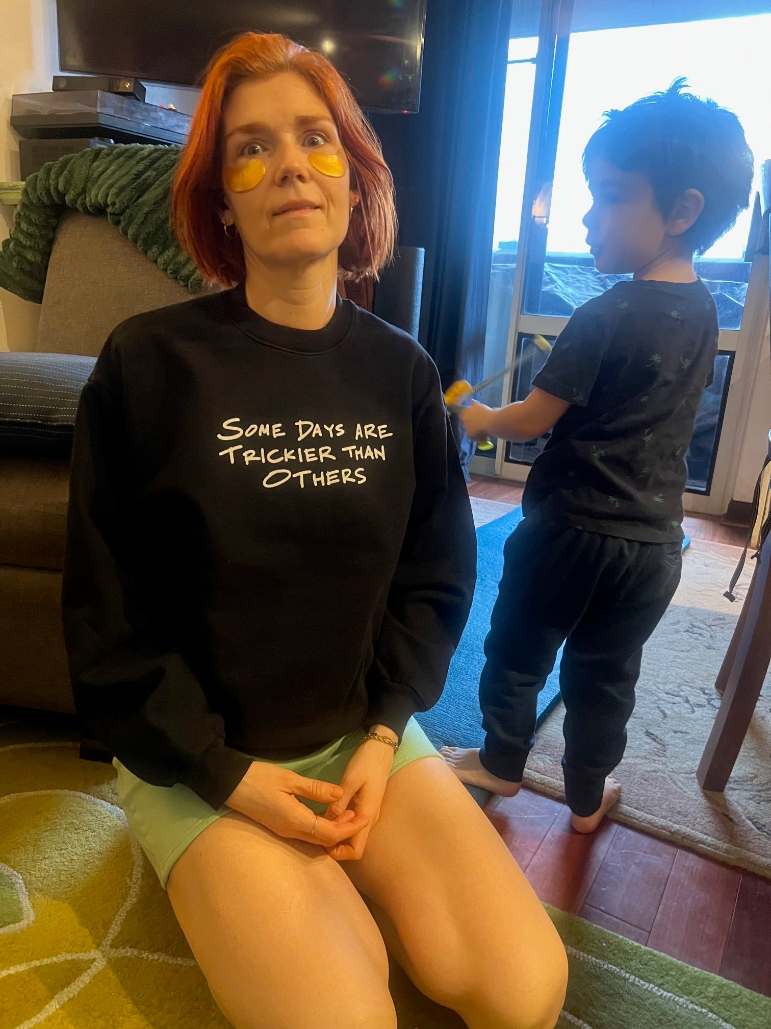 Woman wearing a black sweatshirt with text, sitting on the floor with a child in the background.