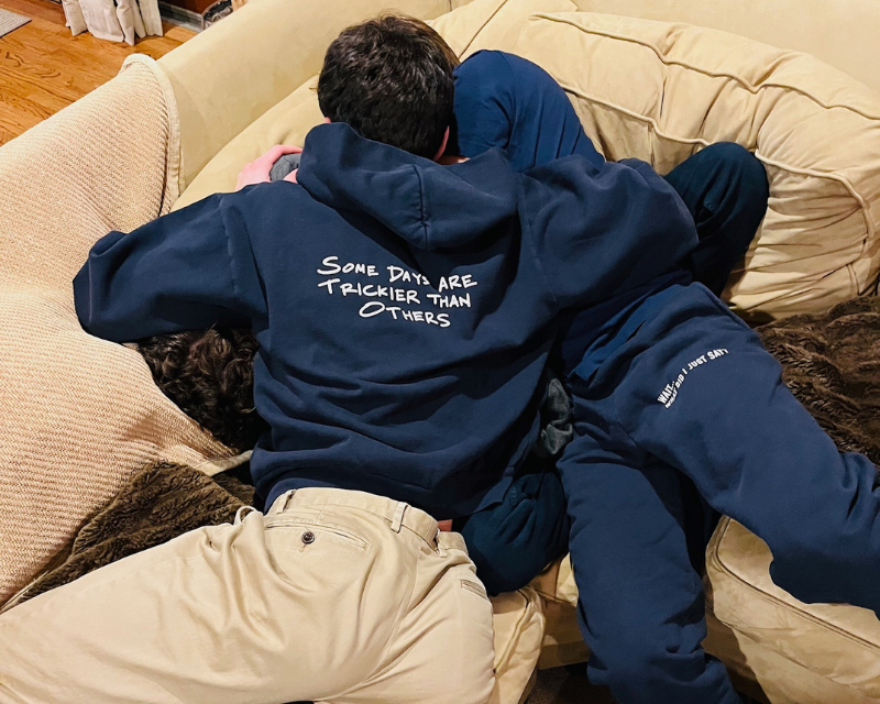 Two people wearing a navy blue sweats with text on the back, sitting on a beige couch.