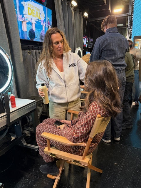 Person sitting in a chair with another person standing next to them, in a backstage or studio setting.